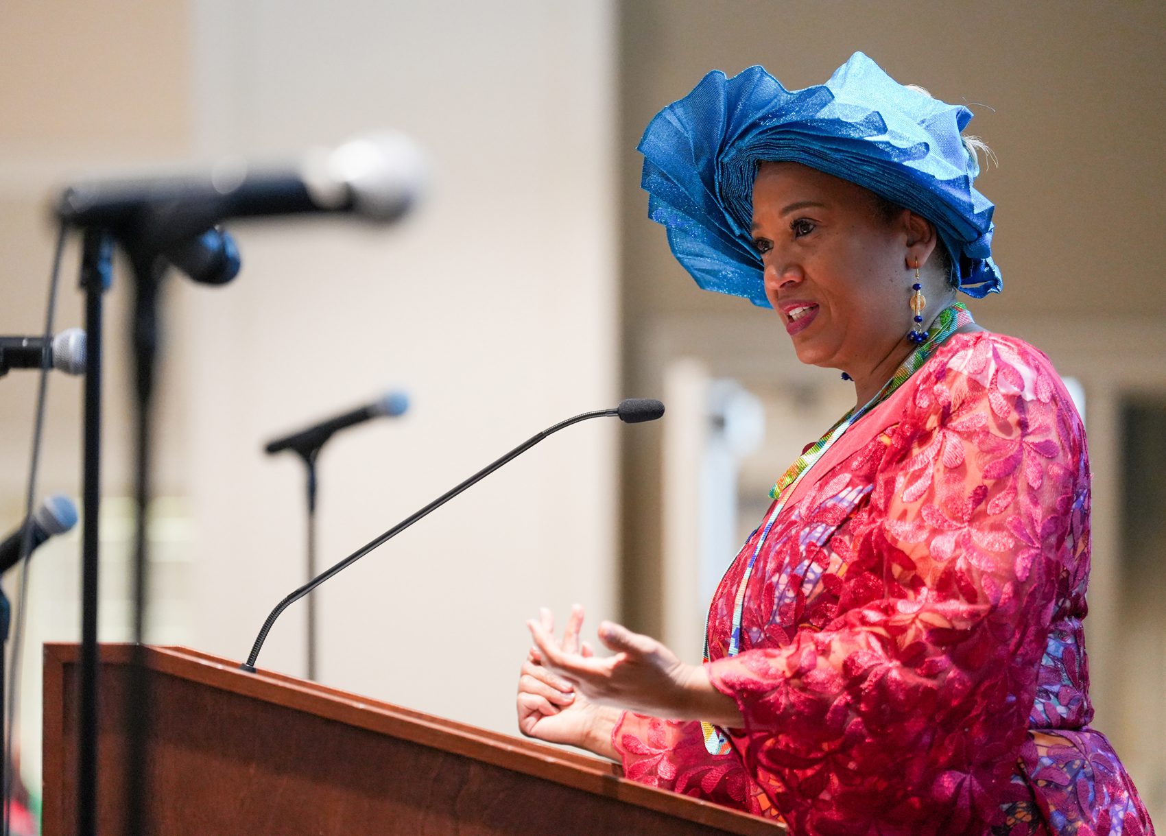 A Black woman in a blue hat and pink dress stands behind a lectern speaking into a microphone.