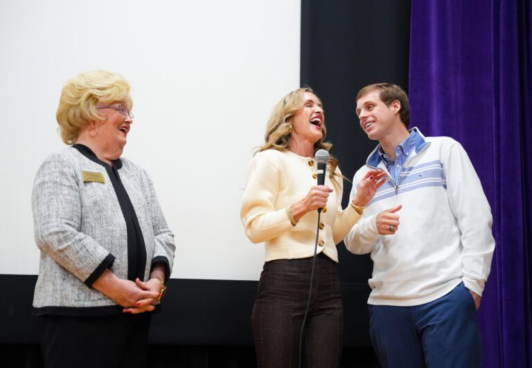 Three people stand on a stage, two of them speaking into a microphone while the third laughs.