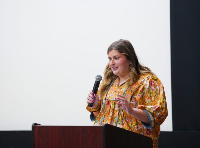 A young woman holding a microphone stands behind a lectern and speaks to an unseen crowd.