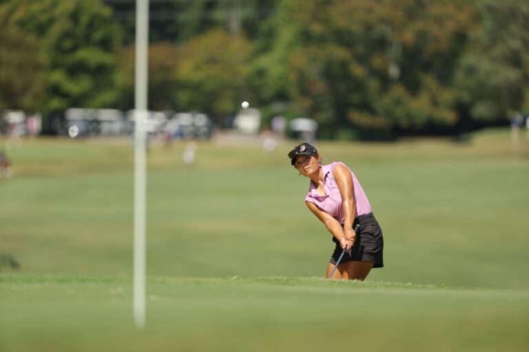 A young woman in a pink polo holds a golf club as she watches the ball soar toward the putting green during a round of golf.