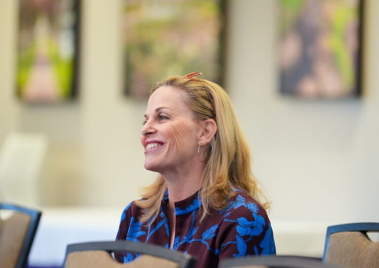 A white woman smiles indoors as she looks at something offscreen