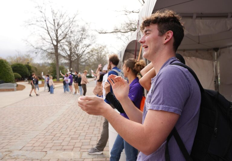 A young man wearing a backpack claps for a group of younger students amid a crowd of other celebrants