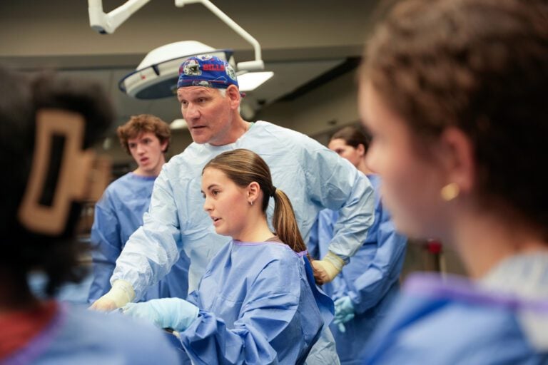 A surgeon wearing a surgical gown and cap helps younger students perform surgical techniques in a teaching lab.