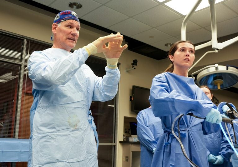 A young woman manipulates surgical instruments while a surgeon beside her gestures with his hands to help guide her movements.