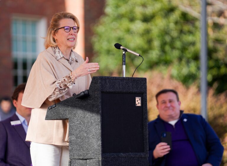 A white woman at a lectern introduces a man seated behind her.