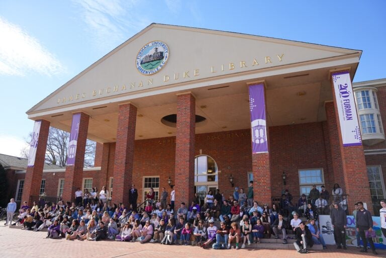 A large crowd of young people are seated on the steps of a university library building