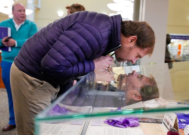 A white man is bent over a glass display case looking at a photo, his face reflected perfectly in the glass as if he is looking at himself.
