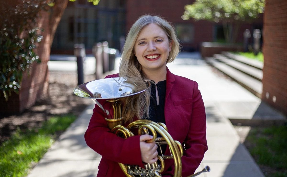 A white woman with blond hair holds a French horn in an outdoor photo. Anna Eberwine