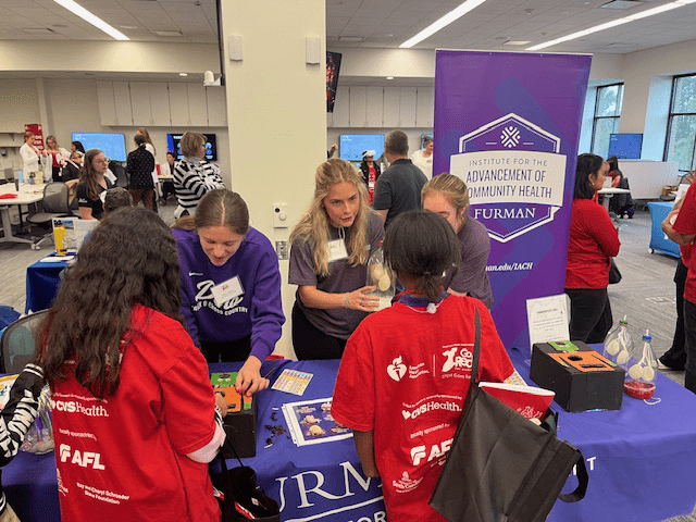 Three college-aged women in purple shirts speak to two middle school students speak to them at a table during an indoor event.