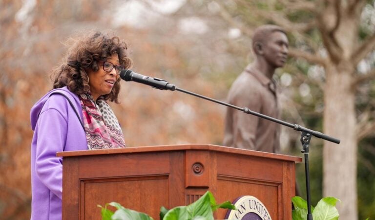 A Black woman wears light purple and speaks at a podium in an outdoor plaza. A bronze statue is in the background. Lillian Brock Flemming