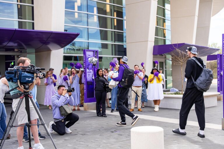 Men's basketball players walk into a building as people cheer them on, holding pom-poms as photographers capture images of the festivities.