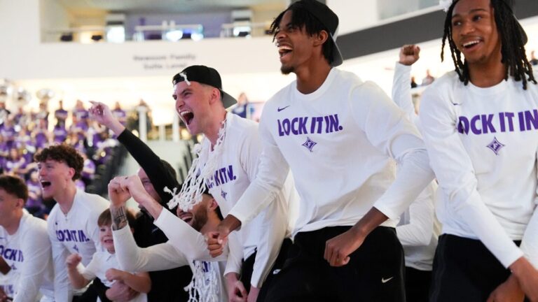 A men's basketball team in white sweatshirts cheer ans celebrate alongside a crowd at an indoor event.