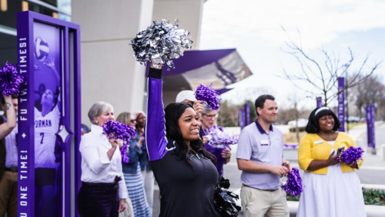 A Black woman cheers holding pom-poms, as a crowd behind her does the same.