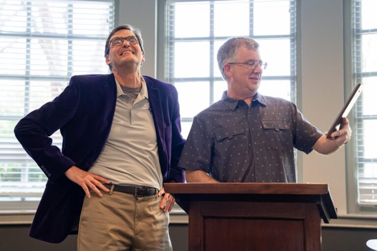 One man stands holding a plaque as he presents an award to another man beside him, who is laughing with his hands on his hips.
