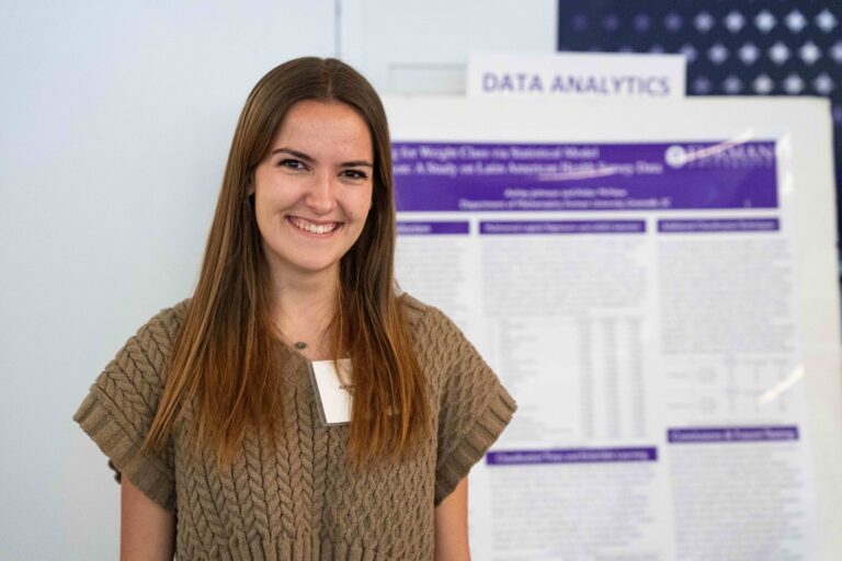 A portrait of a woman smiling and standing in front of a display board showing research related to data analytics.