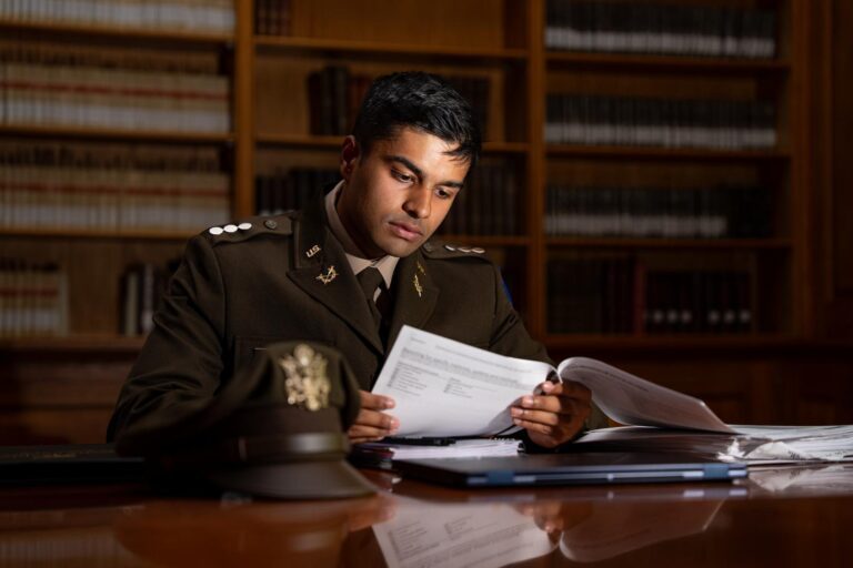 A young man in a military ROTC uniform sits at a table while reviewing documents inside of a library.