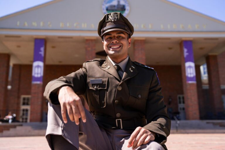 A young man in a military uniform sits outdoors in front of a university library.