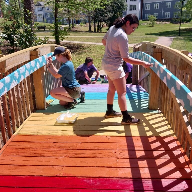 A group of young people paint a wooden bridge outdoors.
