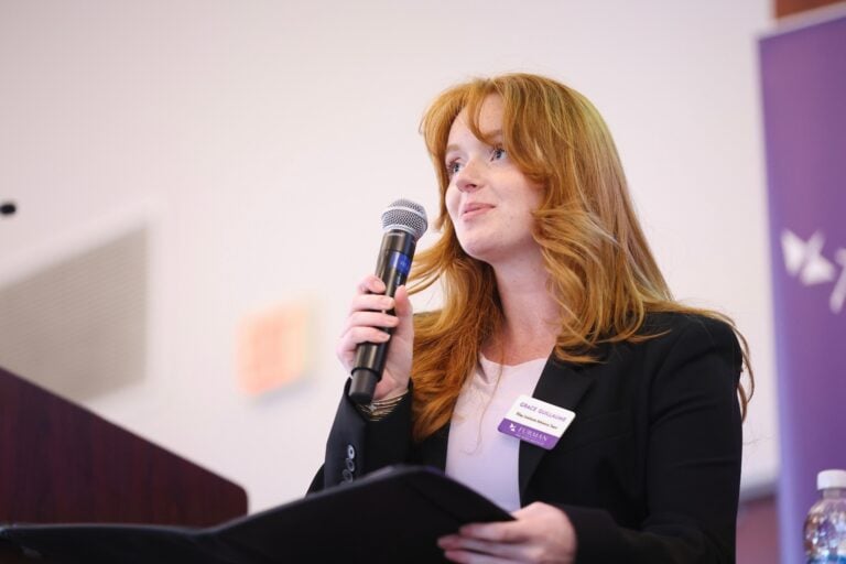 A young woman wearing business attire holds a microphone as she speaks to an unseen crowd.