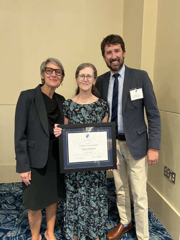 A vertical image of three people posing for an image, with the woman in the middle holding a framed award certificate.