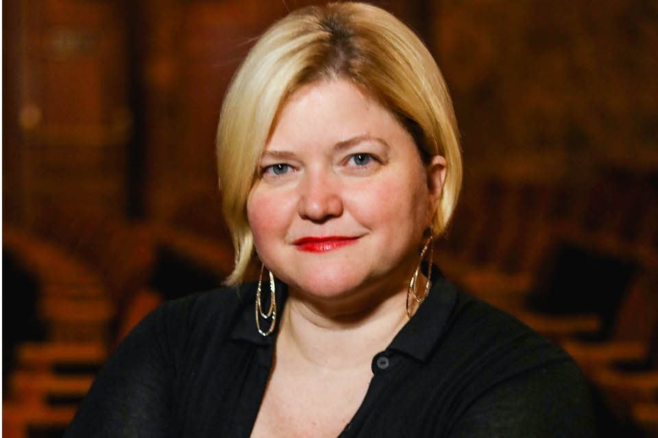 A portrait of a white woman wearing a black shirt with gold earrings and an ear-length haircut.
