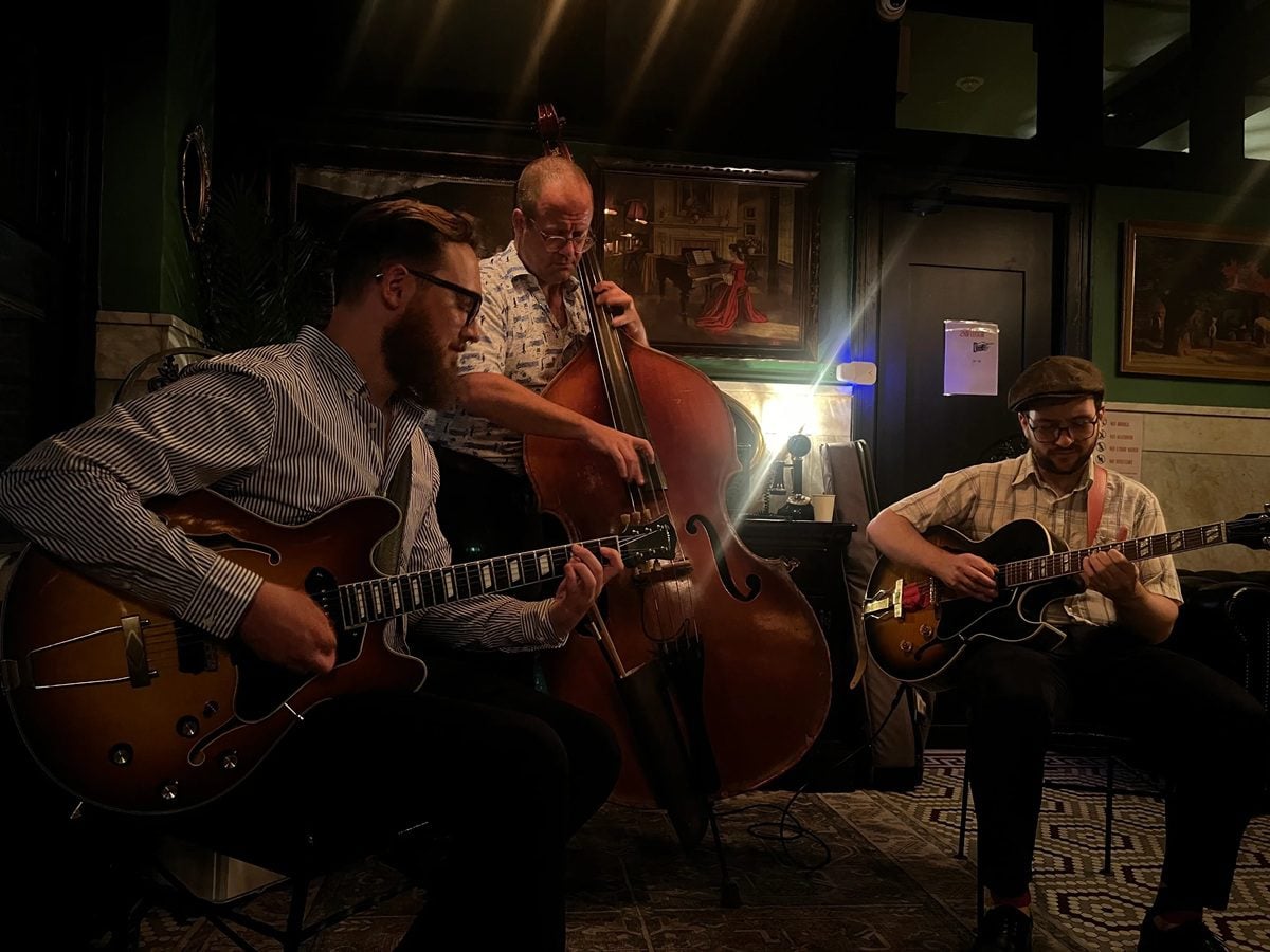 A musical trio of four white men playing jazz guitar and base. Straight Up!