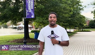 A Black mad in a white T-shirt holds a microphone on a college campus.