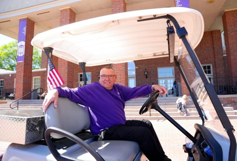 A man smiles at the viewer from behind the steering wheel of a golf cart parked in front of a brick building.
