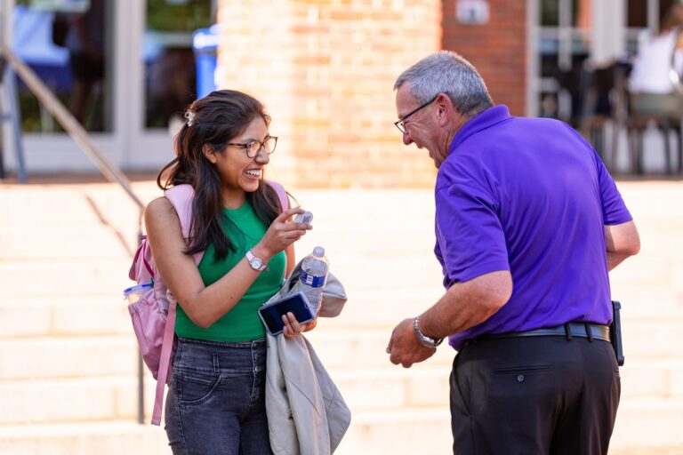 A young woman looks at a sticker she just received from a man, and the two share a laugh in front of the stairs to a brick building.