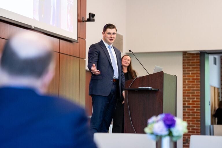 A photo of a young man in a suit jacket, standing and speaking from behind a lectern and gesturing to a seated man in the foreground.