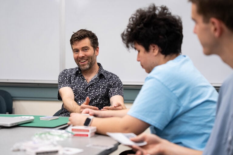 A professor is seated and talks to seated students who have decks of cards and other items used in magic tricks on a desk in front of them.
