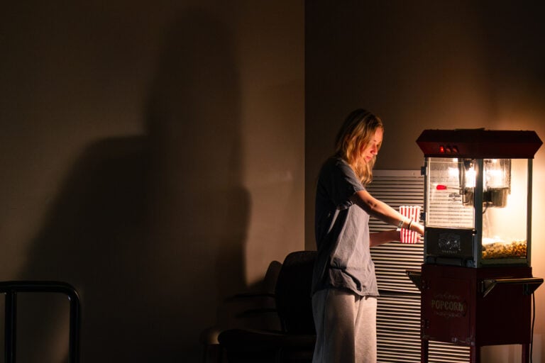 A young woman casts a large shadow on a wall while scooping popcorn out of a lit popcorn machine.