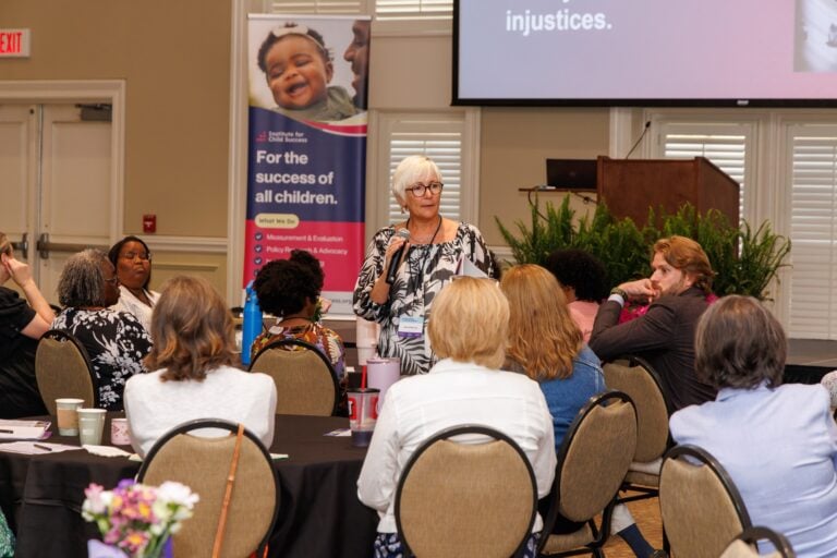 A older woman stands holding a microphone while a crowd of seated people listen to her talk.