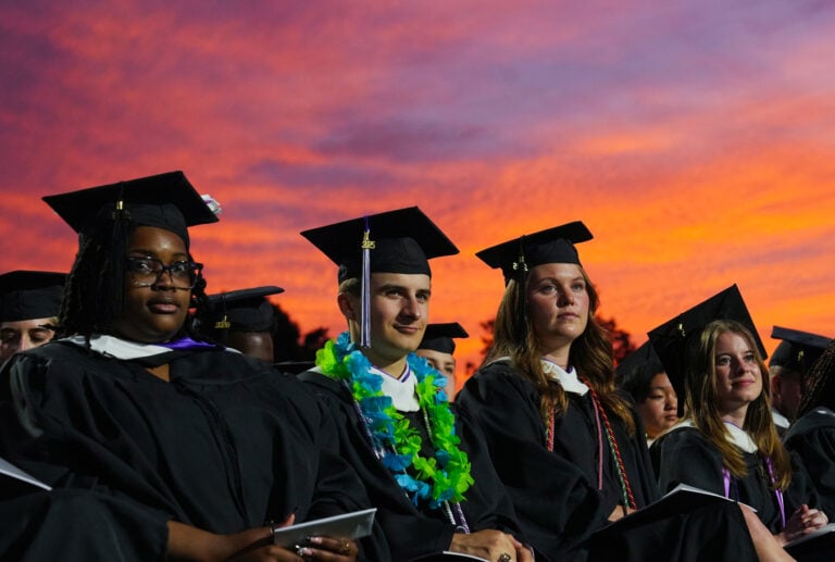 Four students in graduation caps and gowns sit in front of a sunset-tinted sky.