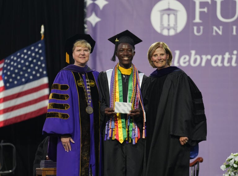 Two women in academic robes stand on either side of a student wearing a graduation cap and gown and holding an award.