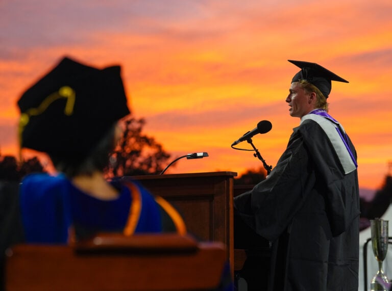 A young man gives a speech from behind a lectern in front of a sunset-colored sky.