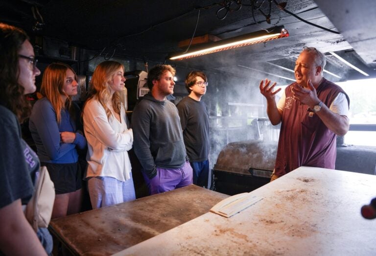 An older man gestures as he speaks with a group of younger people in a barbecue restaurant's smokehouse.