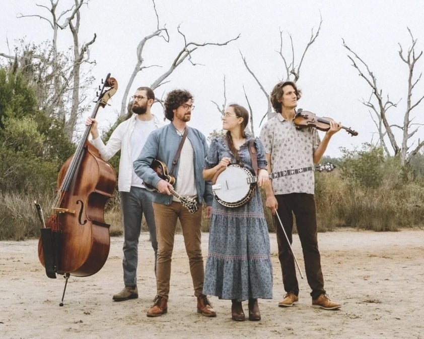 A group of four white band members stand on a beach with leafless trees.