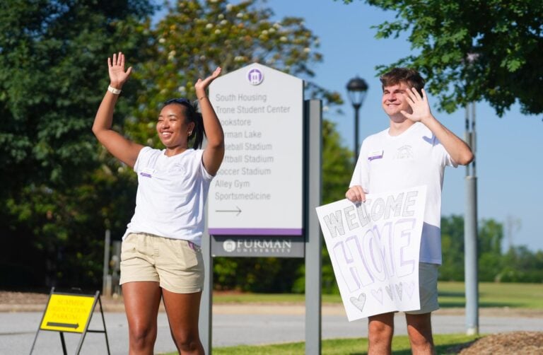 Two Furman students wave at incoming students arriving on campus for orientation, as they hold a sign that reads "welcome home."
