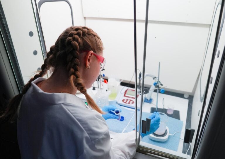 A young woman leans into a fume hood in a laboratory as she mixes a powder into a liquid within a beaker.