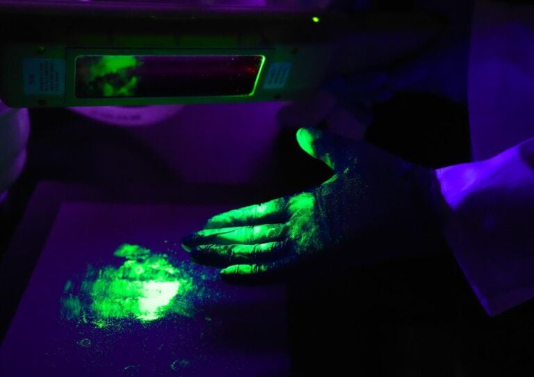 A gloved hand glows neon green under a UV light after touching a nancomposite powder pile.