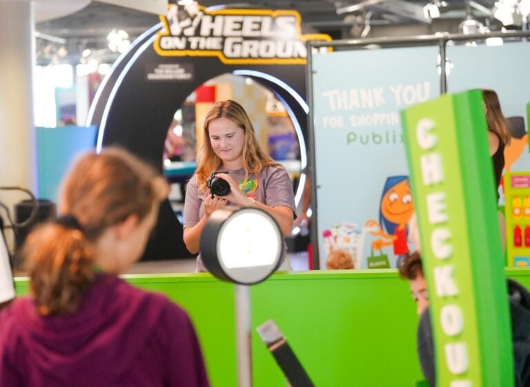 A young woman aims a camera toward children playing in an interactive museum exhibit.