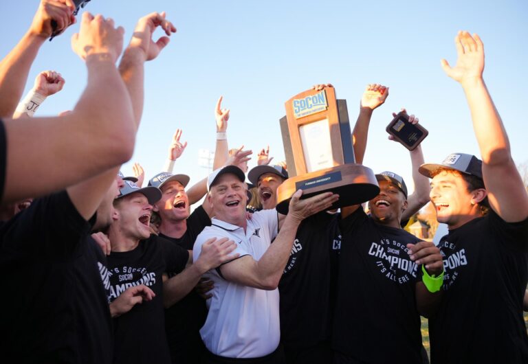 A men's basketball coach and his team cheer and raise their hands in the air as they lift the Southern Conference championship trophy.