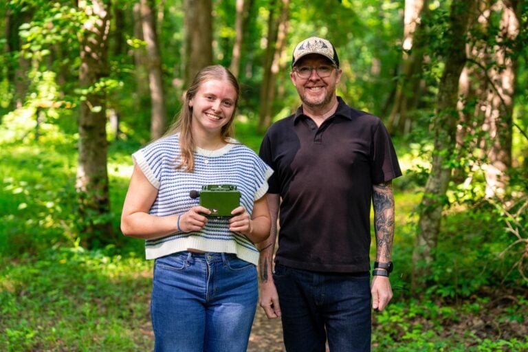 A young woman and her professor pose for a photo in a well-lit wooded area, while the student holds an electronic audio recording device.