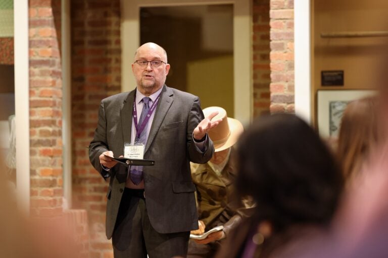 A white man in a suit and tie gestures to people in the foreground as he speaks in an indoor space.