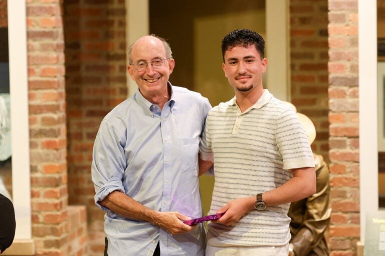 An older white man hands an award to a younger man as they both pose for a photo.