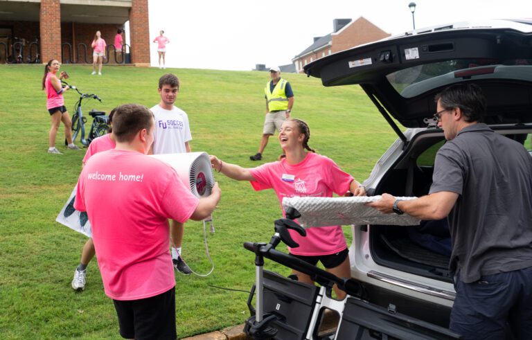 Student orientation helpers in pink shirts unload an SUV and carry items up a hill to a dormitory.