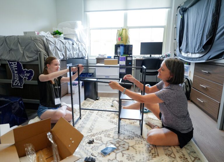 A young woman and her mother work together to assemble a piece of furniture inside a dorm room.