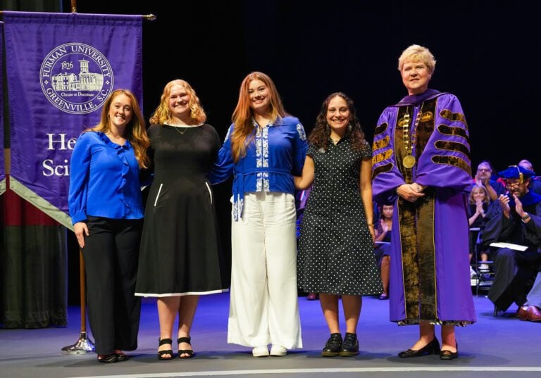 An older woman - a university president - stands beside four students as they pose for a photo.