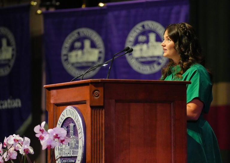 A young woman in a green dress stands behind a lectern and gives a speech with purple banners in the background.